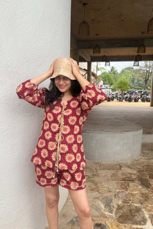 Woman wearing a red floral dress and beige hat standing against a white wall.