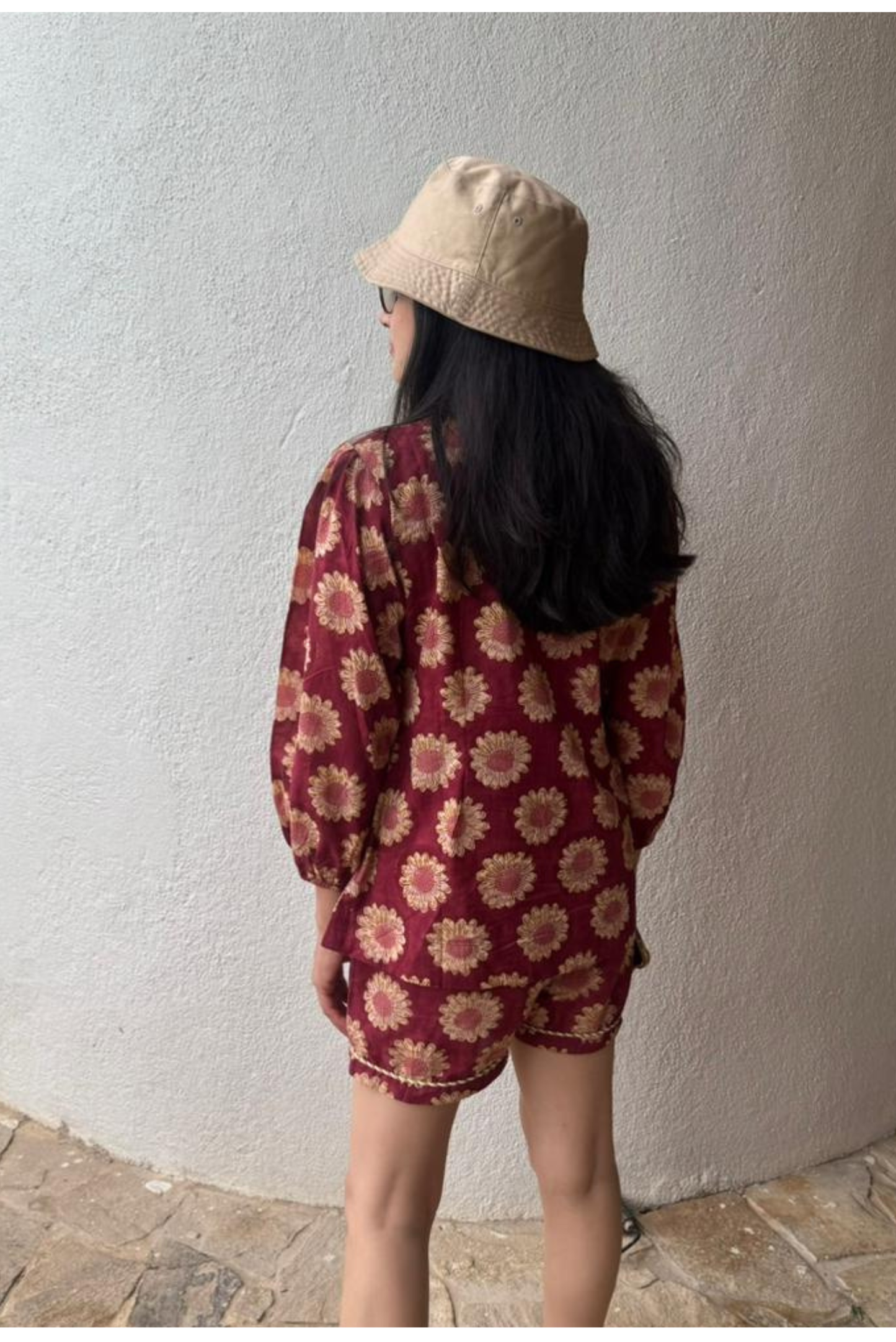 Person wearing a red floral outfit and beige hat against a white wall.